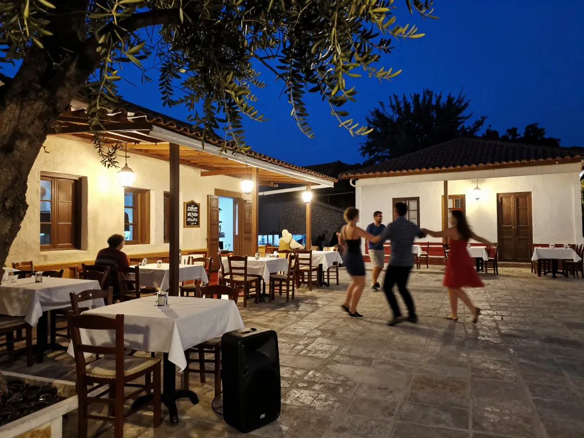 Late-night Greek village square with guests seen from behind starting to dance under lantern light beside tables.