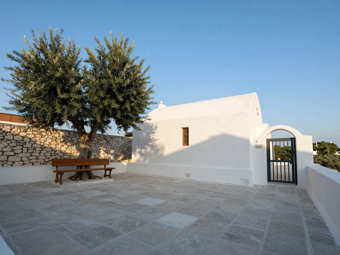 Empty whitewashed chapel courtyard with wooden bench and olive tree, stone walls, blue sky.